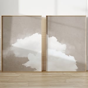 May include: Two framed abstract art prints with a beige background and white cloud-like shapes. The frames are gold.