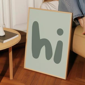 Peut inclure: Une affiche murale encadrée rectangulaire présentant un fond vert sauge clair avec le mot hi écrit dans une grande police arrondie et grasse de couleur vert olive foncé. Le cadre est en bois clair et possède un design simple et épuré.