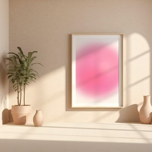 Puede incluir: Una impresi&oacute;n de arte abstracto enmarcada con un dise&ntilde;o degradado en rosa y blanco. La impresi&oacute;n est&aacute; colgada en una pared beige con una planta en maceta y un jarr&oacute;n sobre una superficie blanca.