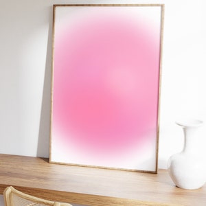 Puede incluir: Una impresi&oacute;n degradada rosa y blanca enmarcada con un marco de madera, sentada en un escritorio de madera con un jarr&oacute;n blanco al lado.