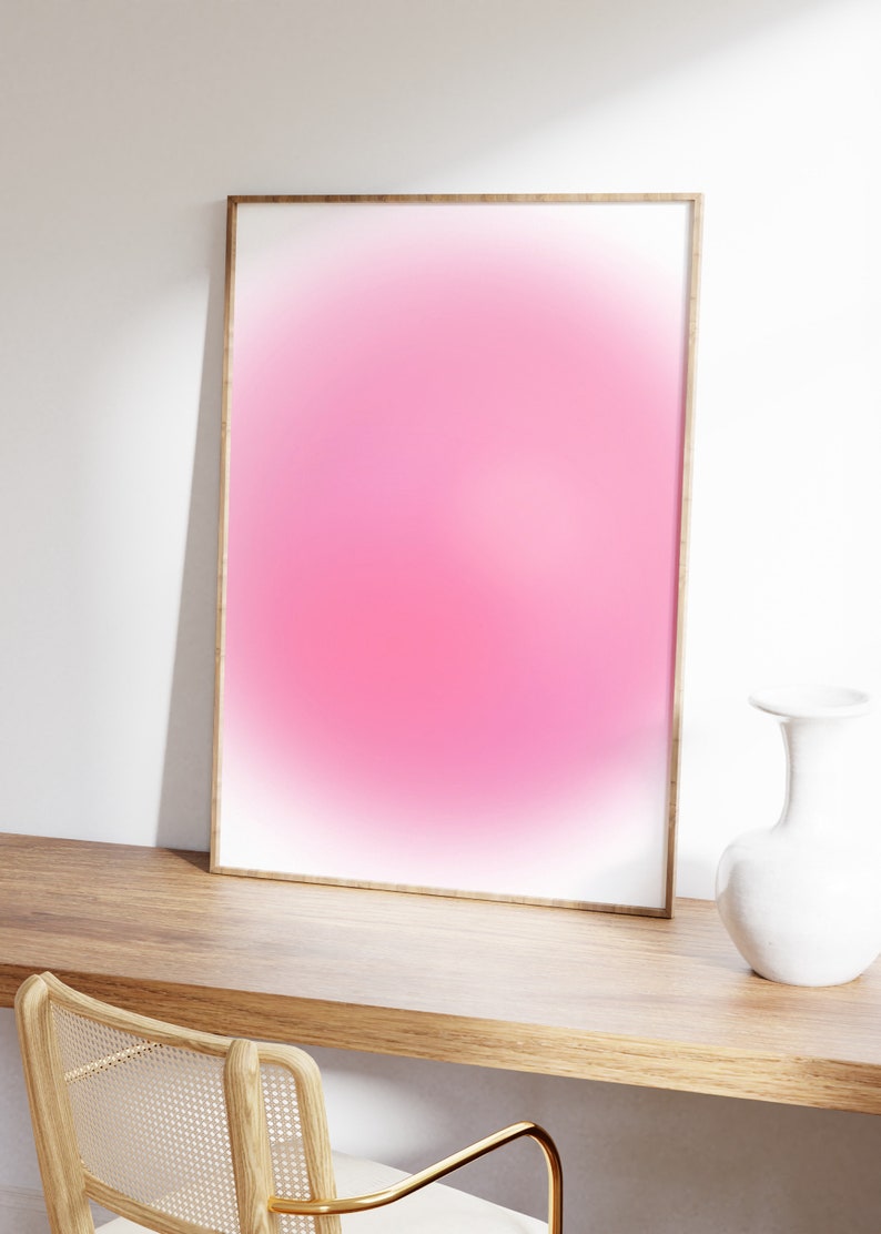 Puede incluir: Una impresi&oacute;n degradada rosa y blanca enmarcada con un marco de madera, sentada en un escritorio de madera con un jarr&oacute;n blanco al lado.