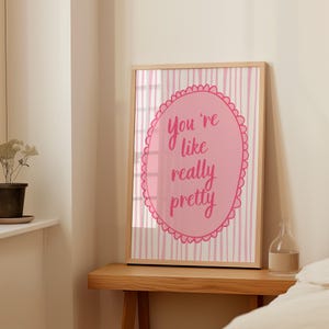 May include: A framed art print with a light wood frame. The print features a pink oval with the words "You're like really pretty" in a handwritten style. The background has pink and white vertical stripes.