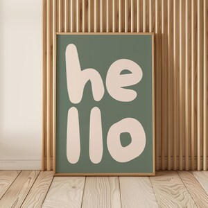 Peut inclure: Une affiche murale rectangulaire dans un cadre en bois clair présentant un fond vert sauge avec le mot hello écrit en grandes lettres arrondies de couleur crème. La typographie est grasse et sans empattement, occupant le centre de l'affiche.
