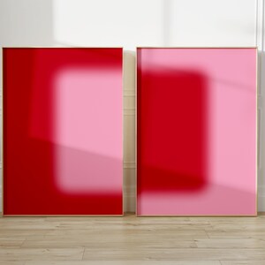 Könnte beinhalten: Zwei gerahmte abstrakte Kunstdrucke. Ein Druck ist hauptsächlich rot mit einem helleren roten Rechteck in der Mitte. Der andere Druck ist rosa mit einem roten Rechteck in der Mitte. Beide Drucke haben goldene Rahmen und stehen auf einem hellen Holzboden.