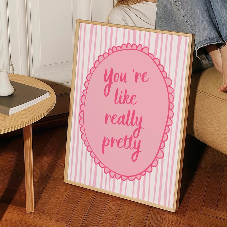 May include: A framed print with a pink and white striped background. The print features a pink oval with the words "You're like really pretty" in a cursive font. The frame is a light wood color.