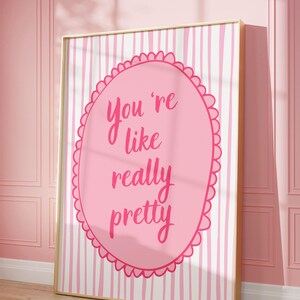May include: A framed pink and white striped print with the text "You're like really pretty" in a pink oval. The artwork is displayed against a pink wall and wooden floor.