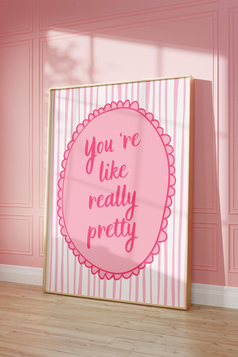 May include: A framed pink and white striped print with the text "You're like really pretty" in a pink oval. The artwork is displayed against a pink wall and wooden floor.