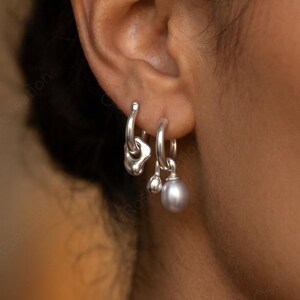 May include: Silver hoop earrings with distinctive organic forms and a dangling pearl. The earrings are worn on the ear of a person with dark skin. The earrings are a stylish accessory.