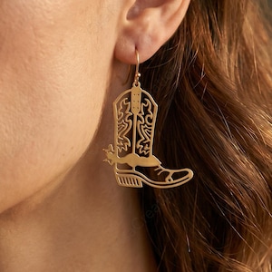 May include: A gold-toned dangle earring shaped like a cowboy boot. The earring features intricate cut-out detailing on the shaft and foot, with a small spur attached to the heel. It hangs from a simple hook wire, showcasing a polished metallic finish.