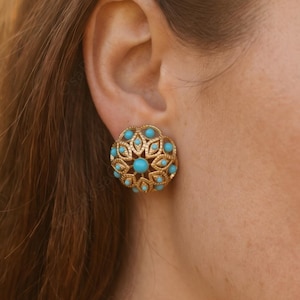 May include: Close-up of a gold-toned stud earring with a circular design. The earring features turquoise-colored stones set within a floral pattern. The earring is on a person's ear.