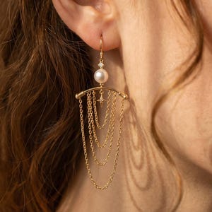 Könnte beinhalten: Ein goldener Hängeohrring mit einer einzelnen weißen Perle, die an einem geschwungenen Stab befestigt ist, sowie vier zarten Goldketten in einem gestuften, kaskadierenden Design. Ein kleiner, vierzackiger Sternanhänger ist mittig am Stab angebracht.