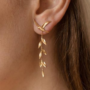 Pode incluir: Brincos dourados com um design de pássaro no lóbulo da orelha, com uma corrente pendurada em forma de folha. Os brincos têm um tom dourado quente, e o design é delicado e detalhado. O fundo é de cor neutra.