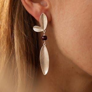 May include: Silver earrings featuring a leaf design. Each earring has a three-leaf cluster at the top, a small dark red bead, and a long, slender leaf shape hanging below. The earrings are worn on the ear.