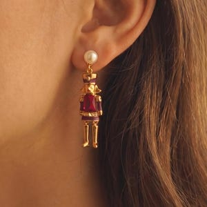 May include: A gold-toned nutcracker earring featuring a small white pearl stud at the top, a deep red rectangular gemstone body, and articulated gold legs. The nutcracker design includes fine gold detailing on the head and torso.