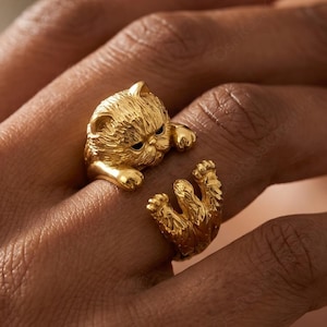 May include: A gold-colored ring in the shape of a cat. The ring wraps around the finger, with the cat's head and paws on opposite sides. The cat's face has detailed features, and the ring has a textured finish.