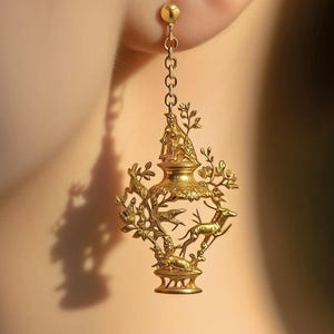 May include: A gold-coloured earring with an intricate design. The earring showcases a central vase-like form, topped with a figure, and surrounded by deer, birds, and foliage. A fine chain links the earring to a small stud.