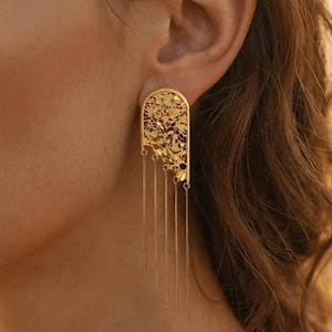 Peut inclure: Boucles d'oreilles dorées avec un motif en filigrane et des brins pendants. Les boucles d'oreilles ont un dessus arqué avec des motifs découpés complexes. Les brins pendants ajoutent du mouvement et une touche d'élégance.