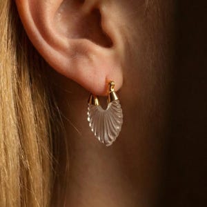 Op de afbeelding: Een close-up van een gouden oorring met een heldere, waaiervormige bedel. De oorring is bevestigd aan een oor, met blond haar op de achtergrond. De bedel heeft een geribbeld ontwerp.
