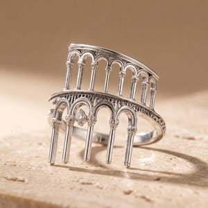 Könnte beinhalten: Ein silberfarbener Ring mit einem strukturellen Design, das eine Reihe von Bogensäulen oder eine Kolonnade nachahmt. Das Metallband ist in zwei Ebenen aus vertikalen Säulen geformt, die durch geschwungene Bögen verbunden sind.