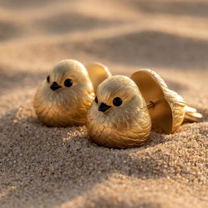May include: Gold-coloured bird-shaped earrings with black eyes and textured feathers. The earrings are displayed on a sandy surface, catching the sunlight. The earrings have a post-back closure.