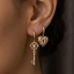 Può includere: Orecchini color oro, uno a forma di chiave e l'altro a forma di cuore con un design a buco della serratura. L'orecchino a chiave ha una parte superiore ornata con una piccola gemma rotonda. L'orecchino a cuore ha una superficie strutturata. Entrambi gli orecchini pendono da piccoli cerchi.
