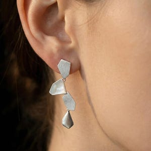 May include: Silver dangle earrings with an abstract design. The earrings feature multiple geometric shapes, including a hexagon and a teardrop, suspended from a post. The earrings are made of a shiny, metallic material.