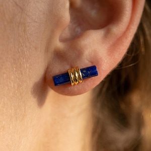 May include: Close-up of a gold and lapis lazuli earring. The earring features a rectangular, deep blue lapis lazuli stone wrapped with three gold bands. The earring is worn on an ear.