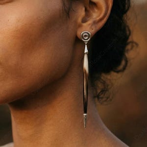 Puede incluir: Pendientes colgantes plateados con un diseño en espiral en la parte superior y un colgante largo y delgado con forma de pluma. El colgante es una mezcla de blanco, marrón y negro. Los pendientes los lleva una persona con cabello y piel oscuros.