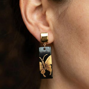 May include: Gold-colored rectangular earrings with a black background and a gold butterfly design. The earrings have a rectangular gold post and a small gold connector. The butterfly is detailed with intricate wing patterns.