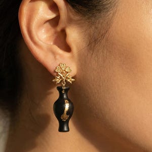 May include: Gold-coloured earrings with a black vase design and gold accents. The earrings have a floral design at the top. The earrings are a unique and stylish accessory, worn on the ear.