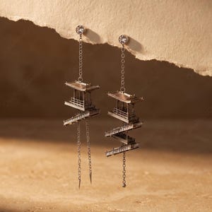 May include: A pair of silver-toned drop earrings featuring a delicate chain attached to a small, clear stud. Each earring displays a miniature, multi-tiered architectural structure resembling a pagoda, connected by thin, dangling silver chains.