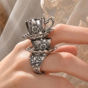 Op de afbeelding: Een zilveren ring met een gestapeld ontwerp van twee miniatuur theekopjes op schoteltjes. Het metalen oppervlak is gedetailleerd met ingewikkelde bloemengravures en bladpatronen op zowel de kopjes als de brede ringband.