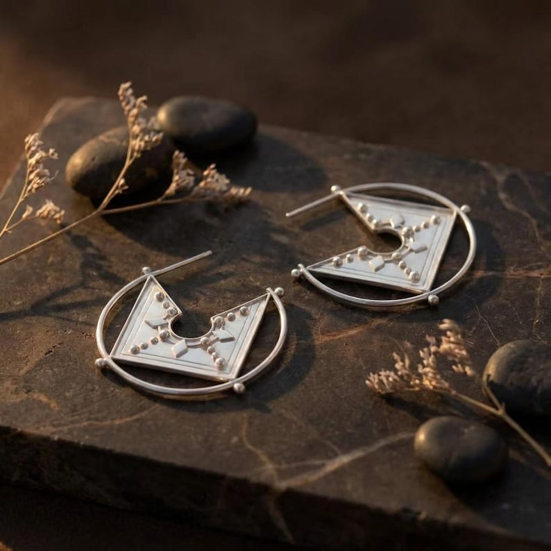 Puede incluir: Un par de pendientes de plata con un dise&ntilde;o geom&eacute;trico. Cada pendiente presenta una forma cuadrada con un recorte y est&aacute; adornado con peque&ntilde;os tachuelas decorativas. Los pendientes est&aacute;n engastados en un marco circular. Joyer&iacute;a.