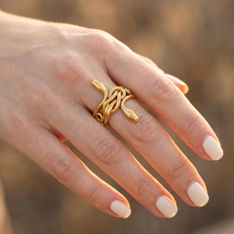 May include: A gold-colored ring featuring a snake design. The ring wraps around the finger with two snake heads facing opposite directions. The snakes are intertwined, creating a unique and eye-catching piece of jewelry.