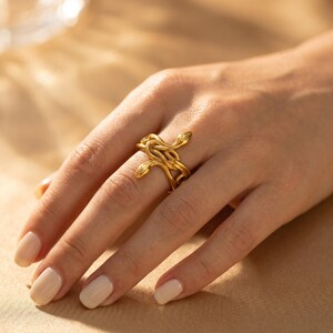 May include: A gold-colored ring featuring a snake design. The ring has a detailed, intertwined snake pattern, with the snake heads visible. The ring is worn on a hand with manicured nails, set against a soft, neutral background.