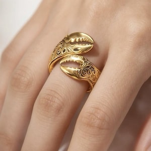 May include: A gold-colored ring shaped like a crab claw, with intricate floral patterns etched into the surface. The ring is worn on a finger, showcasing the detailed design and the way the claw wraps around the finger.
