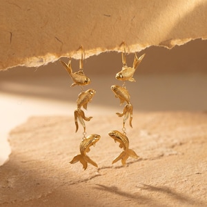 May include: Gold-coloured fish earrings. Each earring features three fish charms suspended from a hook. The fish have detailed fins and tails, and are connected by small chains. The earrings are displayed against a neutral background.
