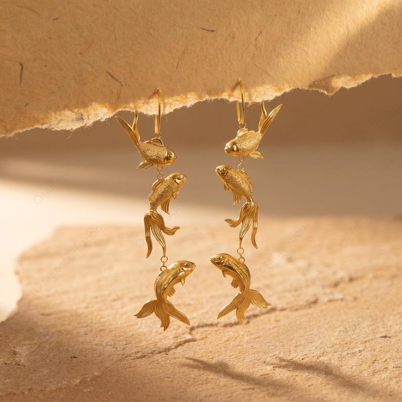 May include: Gold-coloured fish earrings. Each earring features three fish charms suspended from a hook. The fish have detailed fins and tails, and are connected by small chains. The earrings are displayed against a neutral background.