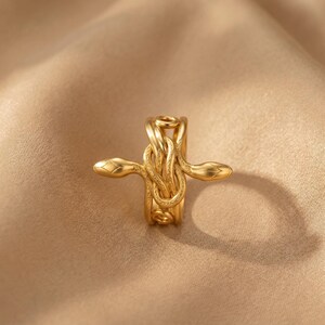 May include: A gold-colored ring featuring a snake design. The ring has two snake heads extending outwards, with the bodies intertwined around the band. The ring is made of a shiny, metallic material.