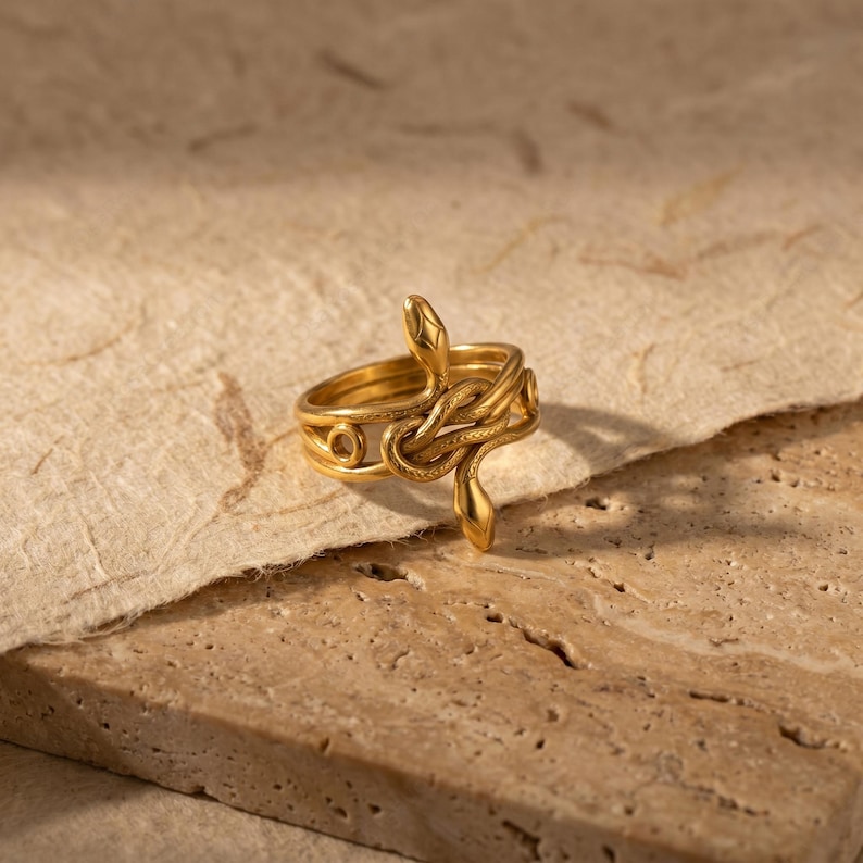 May include: A gold-colored ring with a snake design. The ring features a coiled snake wrapping around the band, with the snake's head and tail extending upwards. The ring is set against a neutral background.