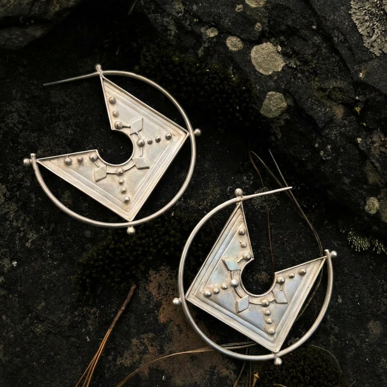 Puede incluir: Un par de pendientes de aro plateados. Cada pendiente presenta un dise&ntilde;o geom&eacute;trico con forma de tri&aacute;ngulo y un marco circular. El tri&aacute;ngulo est&aacute; decorado con peque&ntilde;os puntos en relieve y formas geom&eacute;tricas. Los pendientes se muestran sobre una superficie oscura y texturizada.