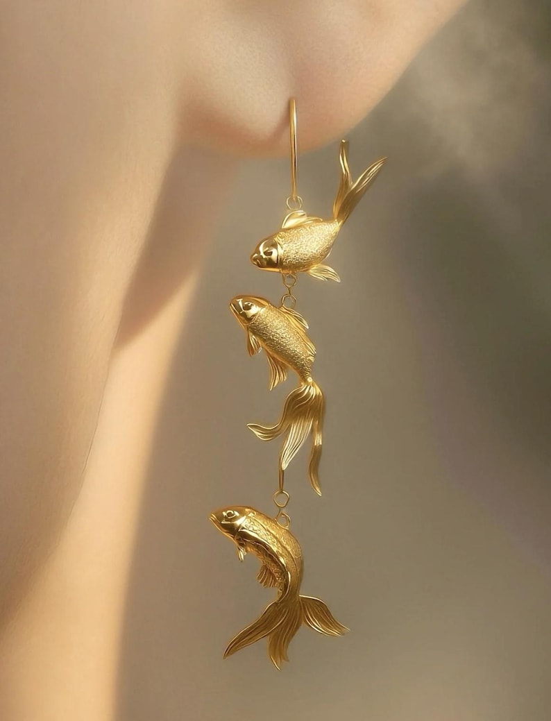 May include: Gold-coloured fish earrings. The earrings feature three detailed fish pendants suspended from a single hook. The fish have textured scales and flowing tail fins, creating a delicate and elegant design. The earrings are designed to be worn.