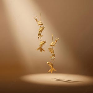May include: Gold-coloured earrings with a cascading design of stylised goldfish. Each earring has multiple fish suspended from a single hook, creating a flowing, aquatic aesthetic. The earrings are set against a warm, neutral background.