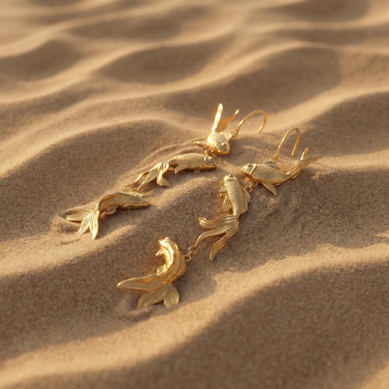 May include: Gold-coloured fish earrings and pendants are arranged on a sandy surface. The earrings and pendants feature detailed fish designs with flowing fins and tails. The jewellery is displayed in a natural setting.