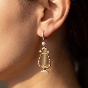 Könnte beinhalten: Goldfarbener Ohrring mit einem Leier-Design, einer Perle und kleinen, klaren Steinen. Der Ohrring hat einen Creolenverschluss mit kleinen, klaren Steinen. Das Leier-Design hat vertikale Linien und eine dekorative Basis.