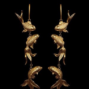 May include: Gold-coloured fish earrings. Each earring features three fish charms suspended from a hoop. The fish have detailed scales and flowing fins. The earrings are set against a black background.