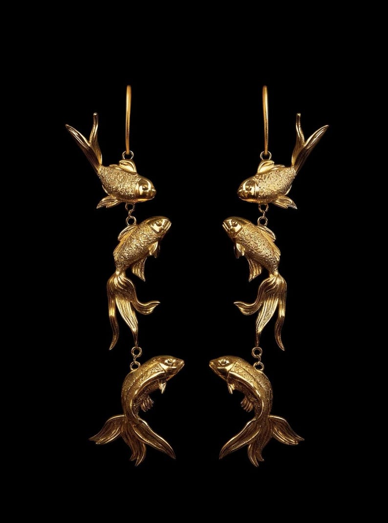 May include: Gold-coloured fish earrings. Each earring features three fish charms suspended from a hoop. The fish have detailed scales and flowing fins. The earrings are set against a black background.