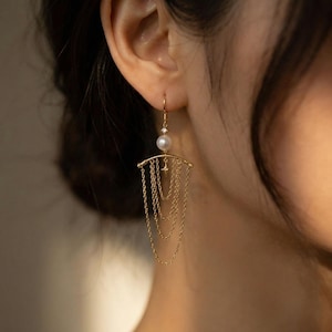 May include: Gold drop earrings with a pearl and a small diamond accent. The earrings have a curved bar design with multiple delicate chain dangles. The chains cascade downwards, creating a layered effect. The earrings are designed to be worn in the ear.