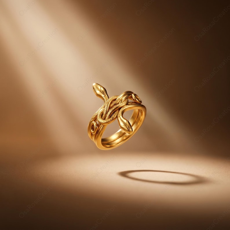 May include: A gold-colored ring featuring a snake design. The ring has two intertwined bands and a snake motif with detailed scales and a raised head. The ring is set against a warm, brown background with a spotlight effect.