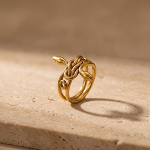 May include: A gold-colored ring featuring a design of intertwined snakes. The ring has a detailed, three-dimensional appearance, with the snakes' heads and bodies forming the ring's structure. The ring is set against a neutral, textured background.